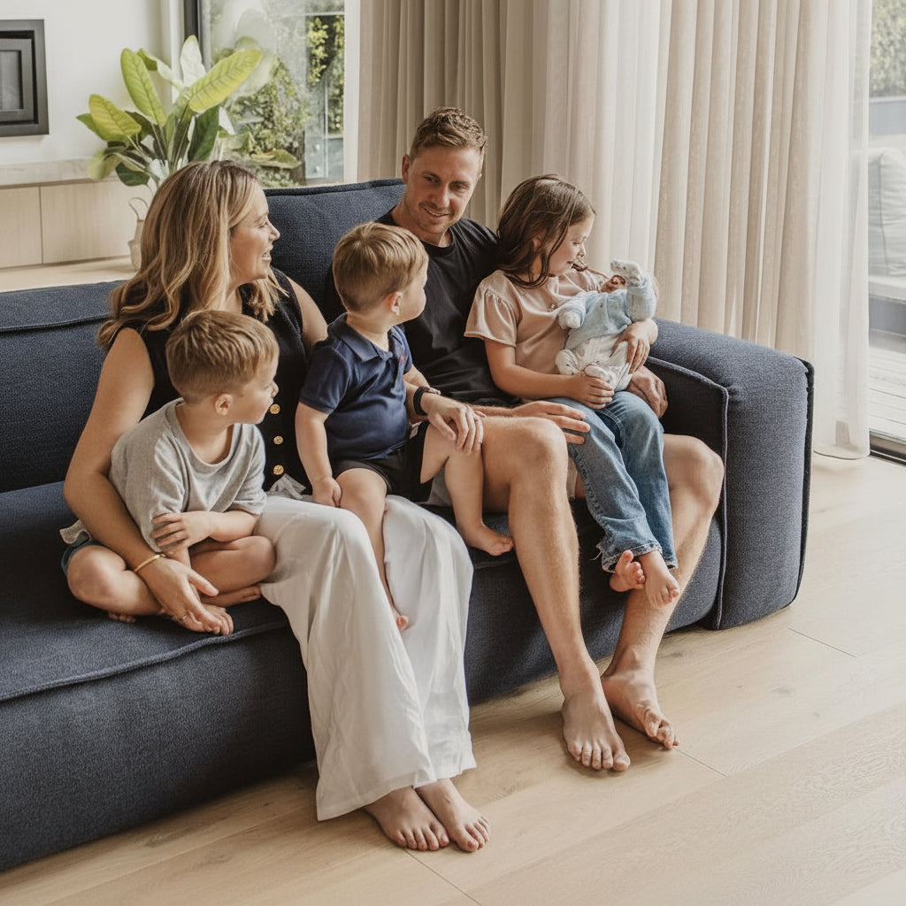 A family of five relaxes on a dark blue sofa in a bright, modern living room with large windows, beige curtains, and a pool deck visible outside.